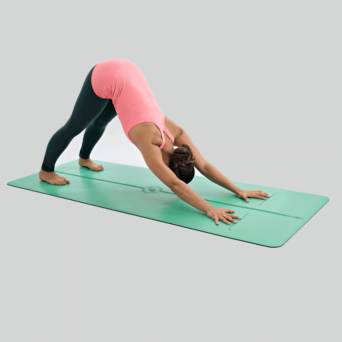A woman performs a downward-facing dog yoga pose on a Liforme Evolve Yoga Mat Green against a clean, light gray studio background.
