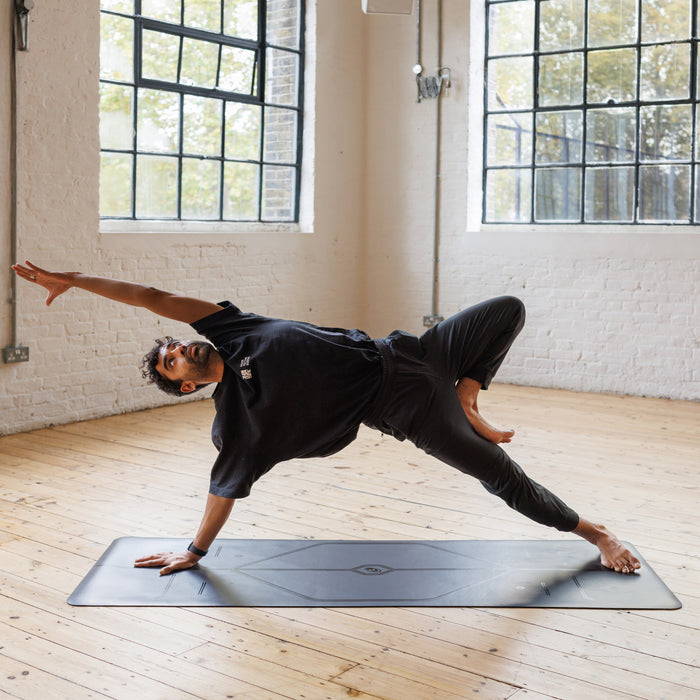 A man performs a side plank yoga pose on a Liforme In Line Black Yoga Mat in a bright yoga studio with wooden floors and large windows.
