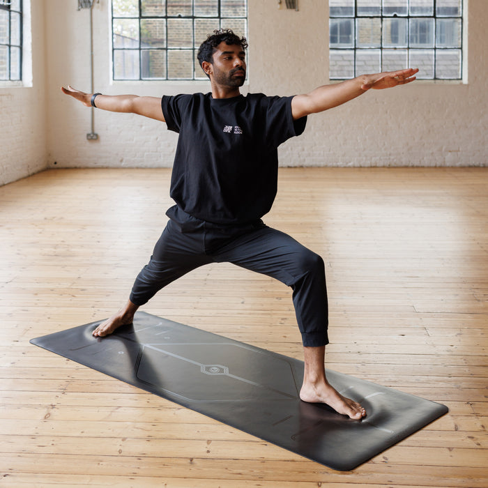 A man performs a warrior II  pose, Virabhadrasana II yoga pose on a Liforme In Line Black Yoga Mat in a bright yoga studio with wooden floors and large windows.