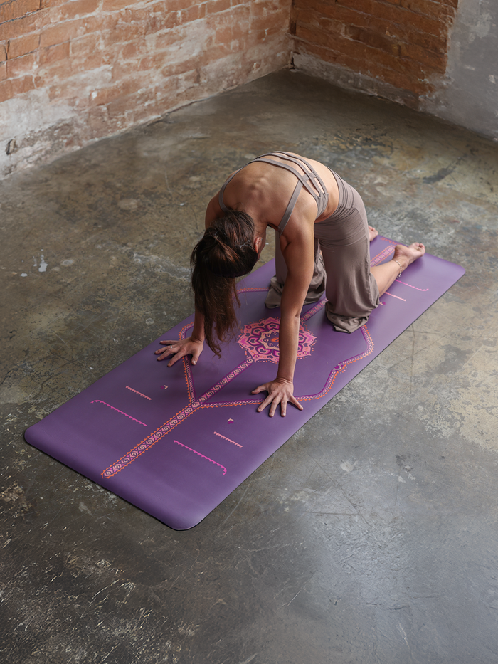 A woman holding a Cat-Cow Stretch on a Liforme Blossoming Lotus Purple Yoga Mat in a yoga studio