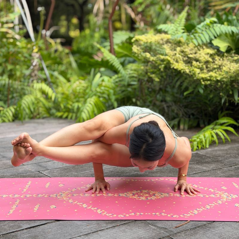 A woman performs a yoga pose on a Liforme Cherry Blossom Pink yoga mat  outdoors on a wooden deck surrounded by lush green plants.
