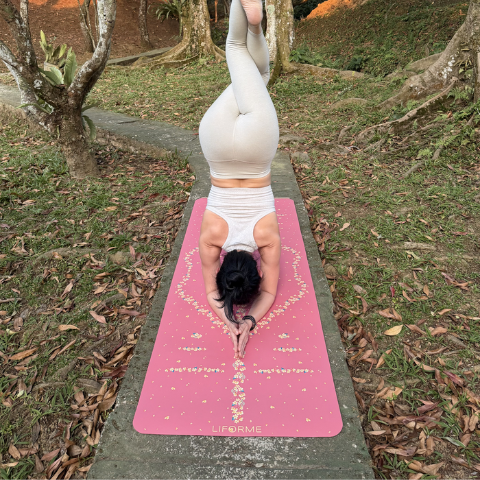 A woman in a headstand yoga pose on a Liforme Cherry Blossom Pink yoga mat outdoors on a concrete path surrounded by grass and trees. 