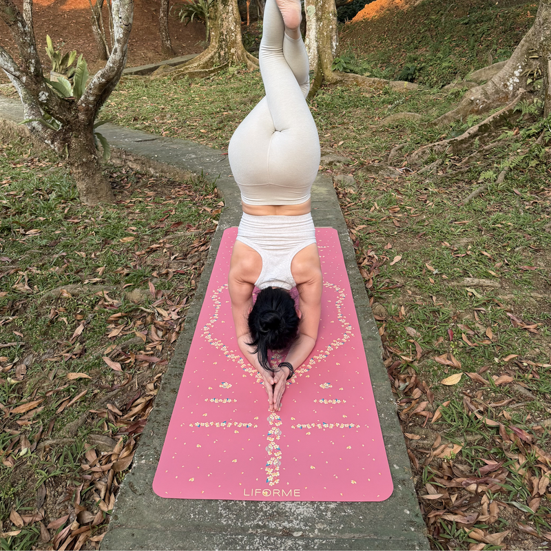 A woman in a headstand yoga pose on a Liforme Cherry Blossom Pink yoga mat outdoors on a concrete path surrounded by grass and trees. 