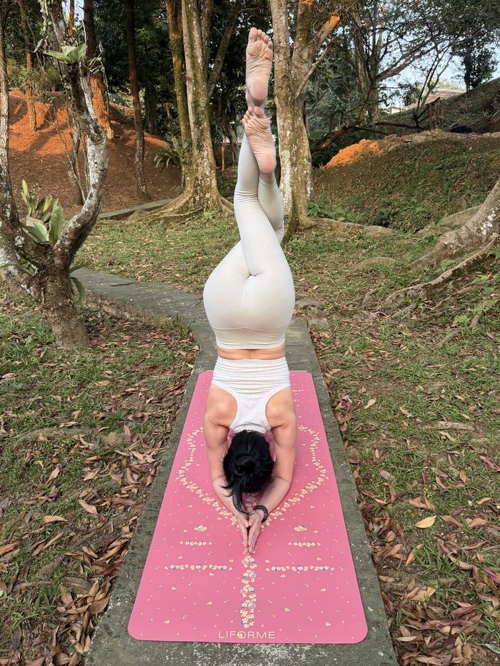 A woman in a headstand yoga pose on a Liforme Cherry Blossom Pink yoga mat outdoors on a concrete path surrounded by grass and trees. 