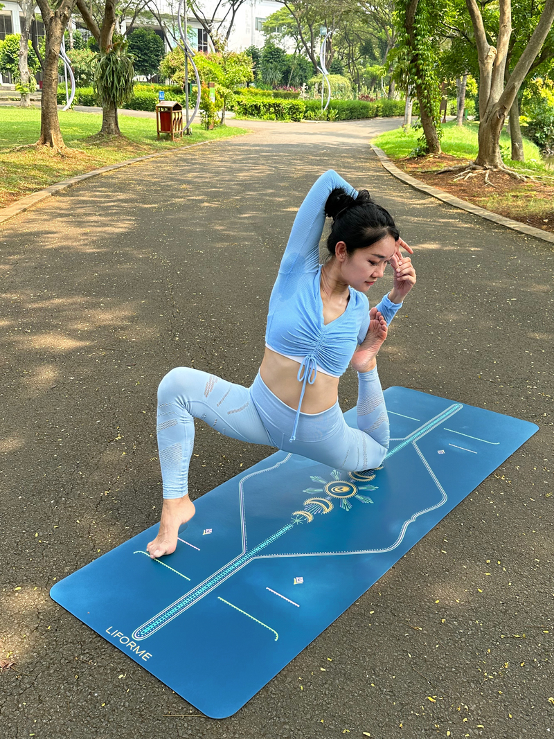 A woman performs a yoga pose on a Liforme Cosmic Moon Blue yoga mat outdoors on a paved park path.