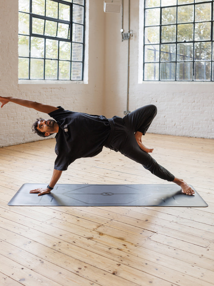 A man performs a side plank yoga pose on a Liforme In Line Black Yoga Mat in a bright yoga studio with wooden floors and large windows.
