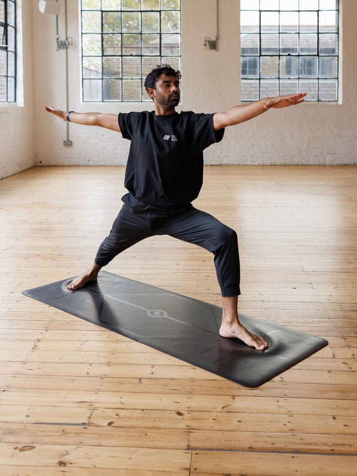 A man performs a warrior II  pose, Virabhadrasana II yoga pose on a Liforme In Line Black Yoga Mat in a bright yoga studio with wooden floors and large windows.