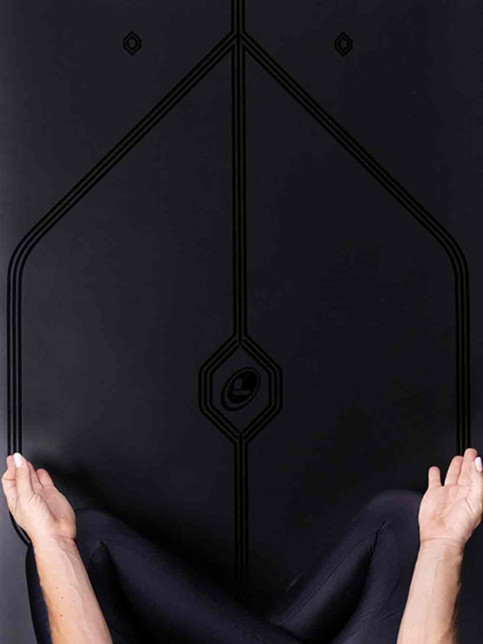 A person sitting on a lotus yoga pose on a Liforme In Line Black Yoga Mat. Showing a close-up of the mat alignment design.