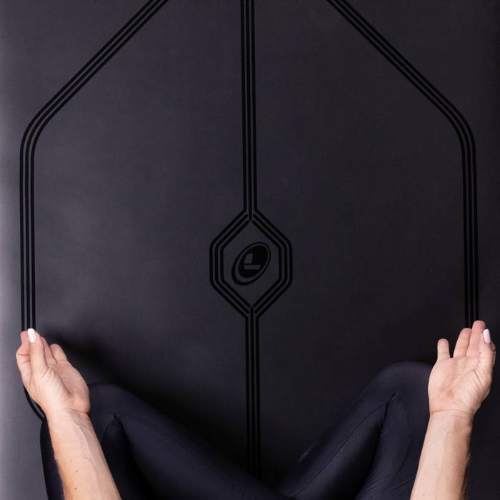 A person sitting on a lotus yoga pose on a Liforme In Line Black Yoga Mat. Showing a close-up of the mat alignment design.