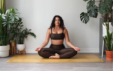Woman in brown athletic wear sitting in a yoga pose on a mat with plants around her.