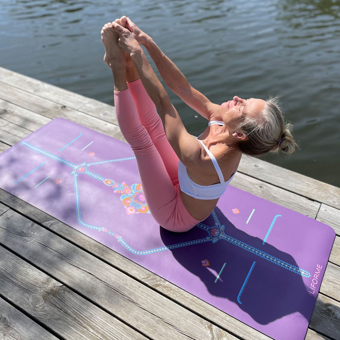 Yogi balancing in Both Big Toe Pose on a Purple Liforme Mindful Garden yoga mat by the water, extending both legs upward while holding the feet for core stability.