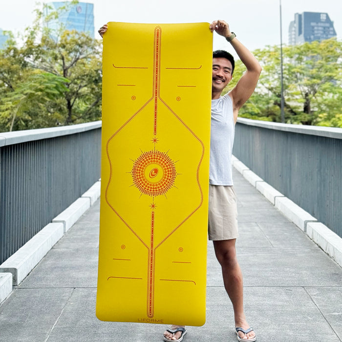Person holding up the Liforme Radian Yoga Mat Yellow to display the full, colourful alignment design in an outdoor setting.