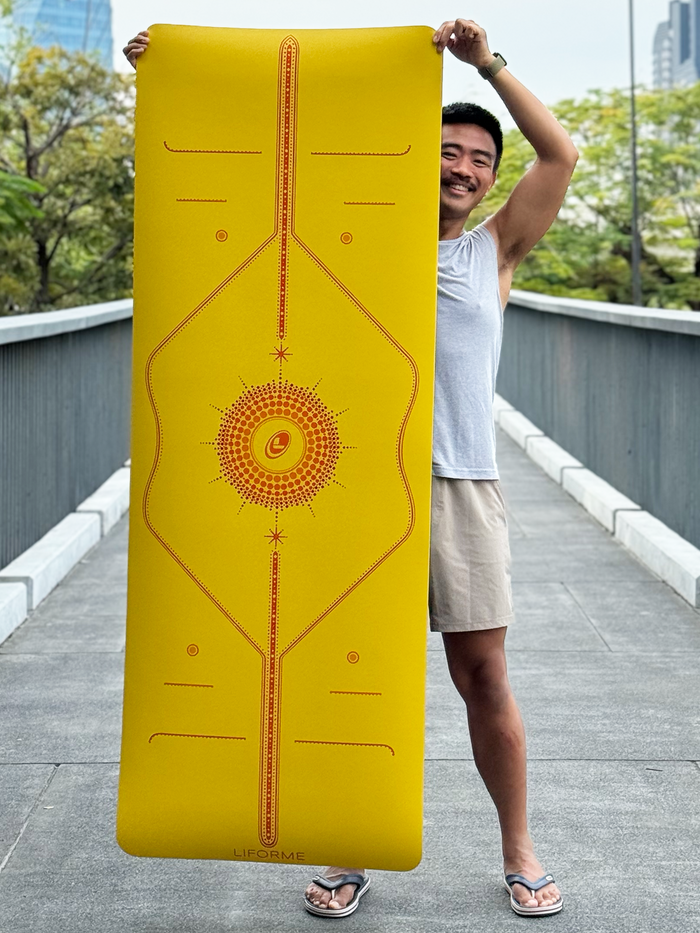 Person holding up the Liforme Radian Yoga Mat Yellow to display the full, colourful alignment design in an outdoor setting.