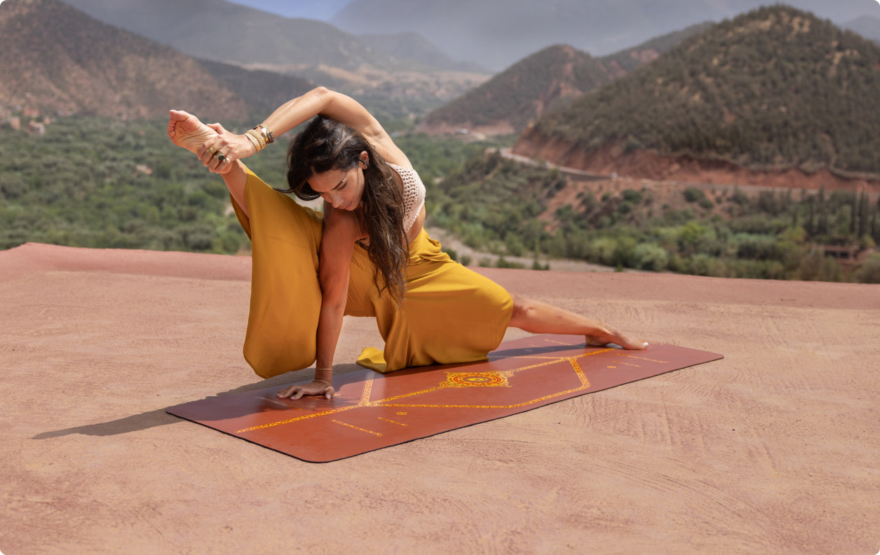 A woman in an advanced side-stretching yoga pose on a terracotta Liforme Yoga Mat with gold alignment guides, situated on an outdoor rooftop with a vast mountain valley background.