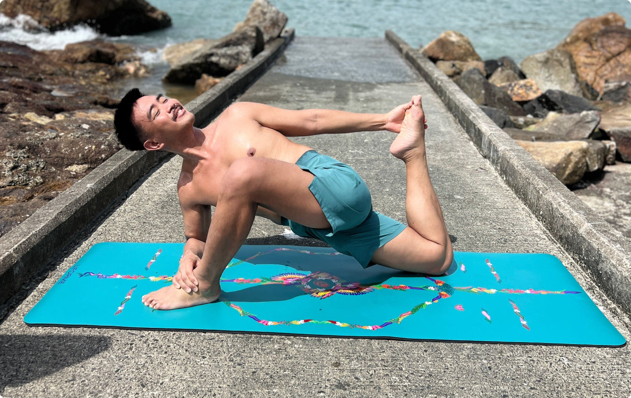 Man practicing a Twisted Dragon/Lizard Pose variation, reaching back to hold his foot, on a teal Liforme Tropical Yoga Mat on a stone coastal walkway.