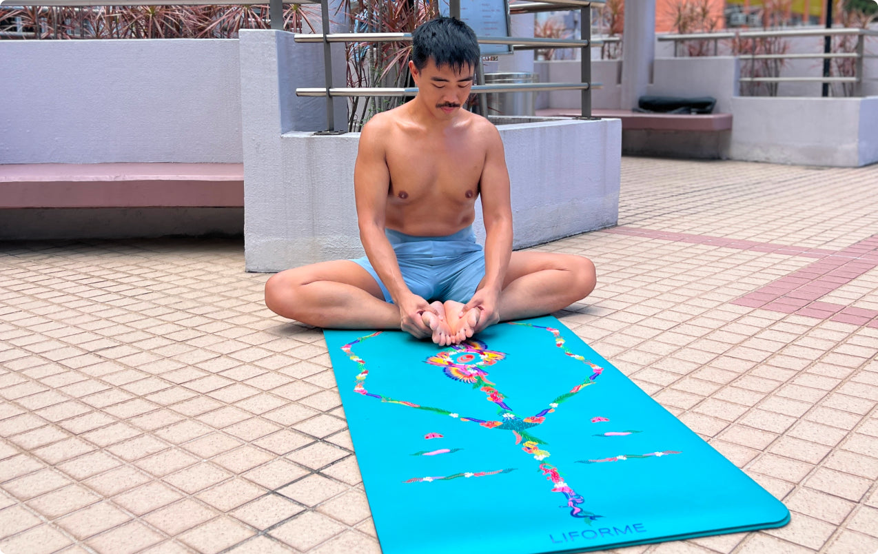 Man sitting in Bound Angle Pose (Baddha Konasana), holding his feet on a teal Liforme Tropical Yoga Mat in an outdoor urban setting.