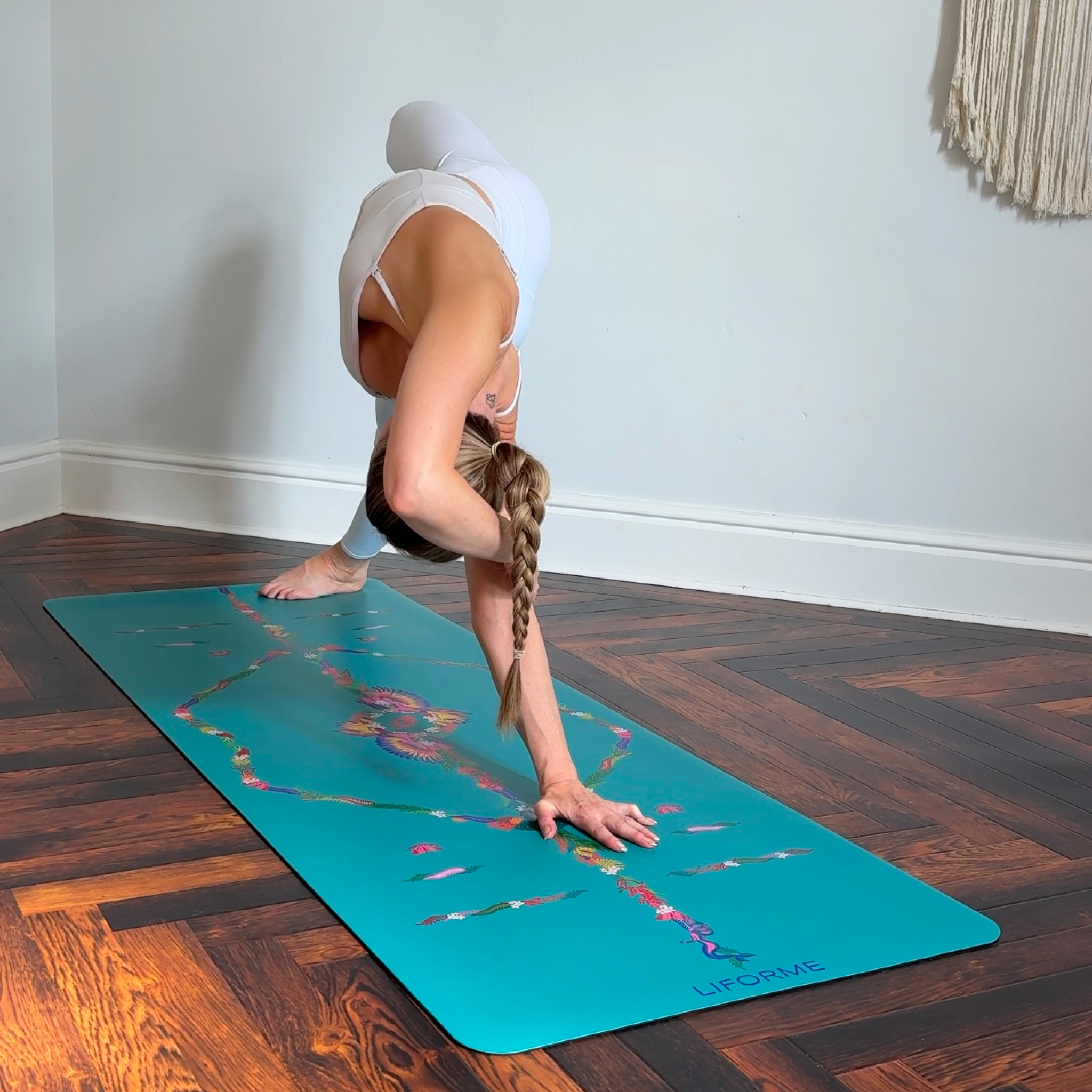 A woman in white activewear performing a Triangular/Twisted stretch variation on a teal Liforme Tropical Yoga Mat on dark wood floors.
