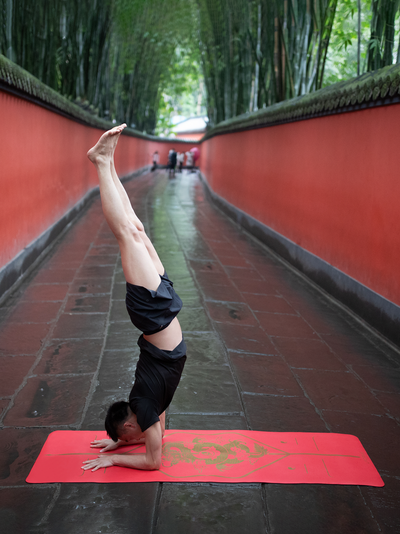 A man performs a headstand on a Liforme Zodiac Dragon, set in a narrow outdoor corridor lined with red walls and green bamboo.